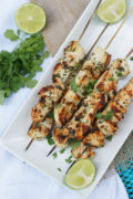 Six garlic chicken skewers on a white plate, garnished with fresh cilantro.