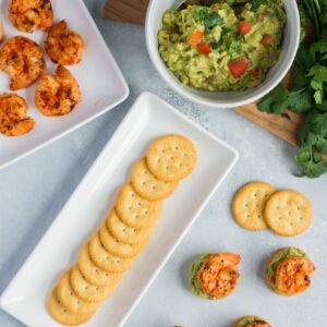 Crackers on a serving plate next to a bowl of guacamole and a plate of shrimp.