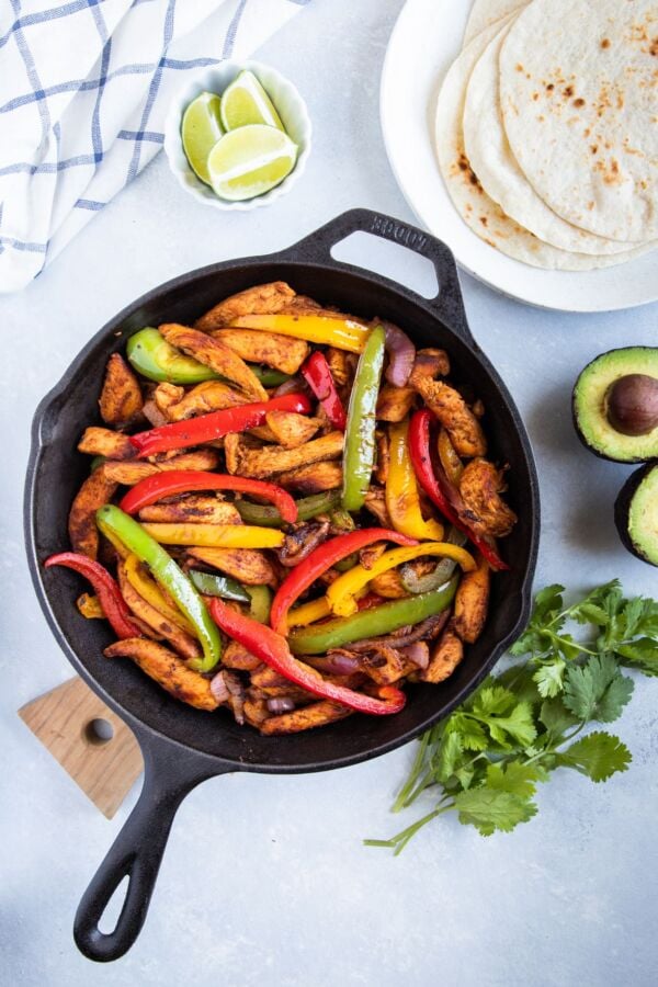 Chicken Fajitas in a skillet with flour tortillas, avocado, cilantro and lime wedges on the side.