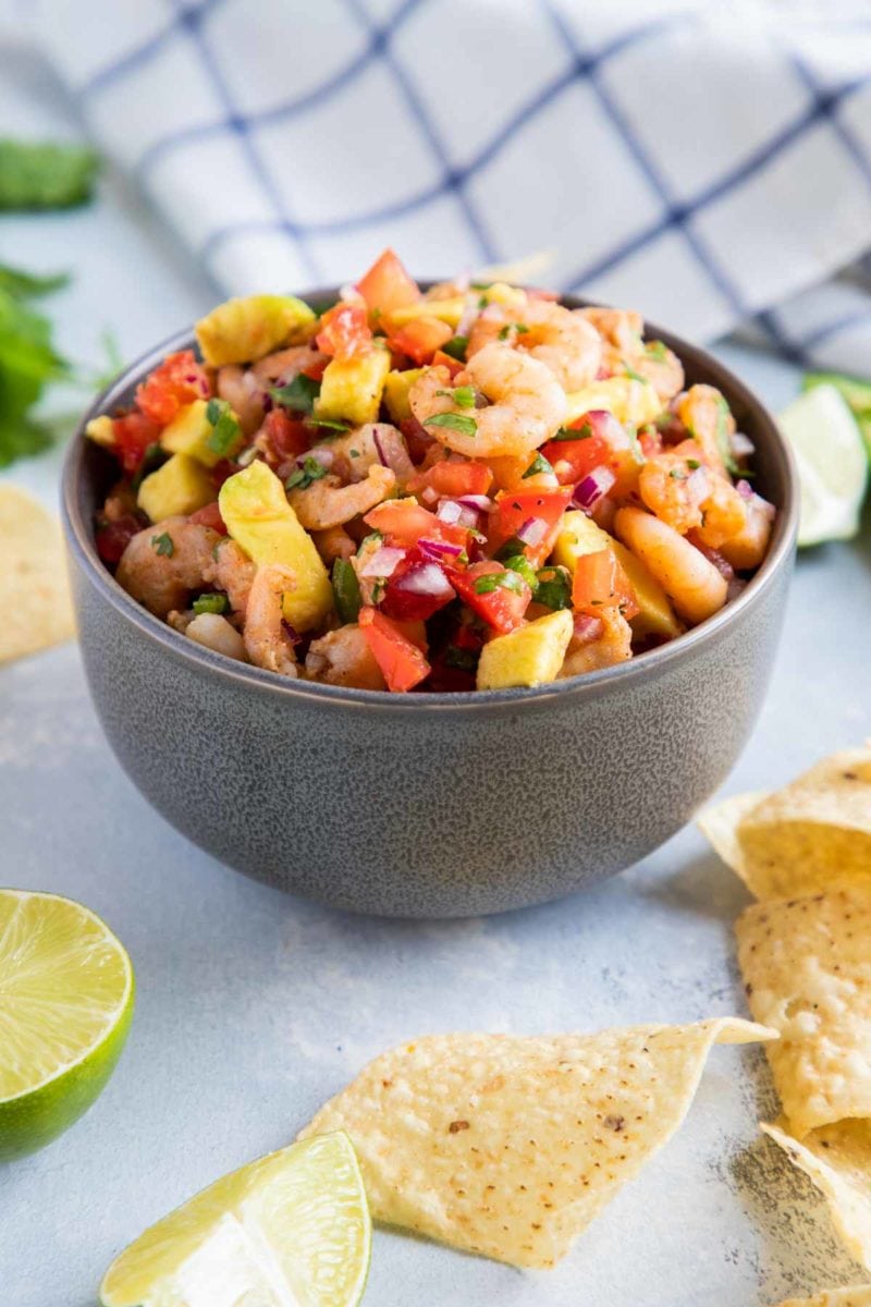 Un tazón lleno de salsa de aguacate y camarones, rodeado de totopos de maíz y rodajas de lima.
