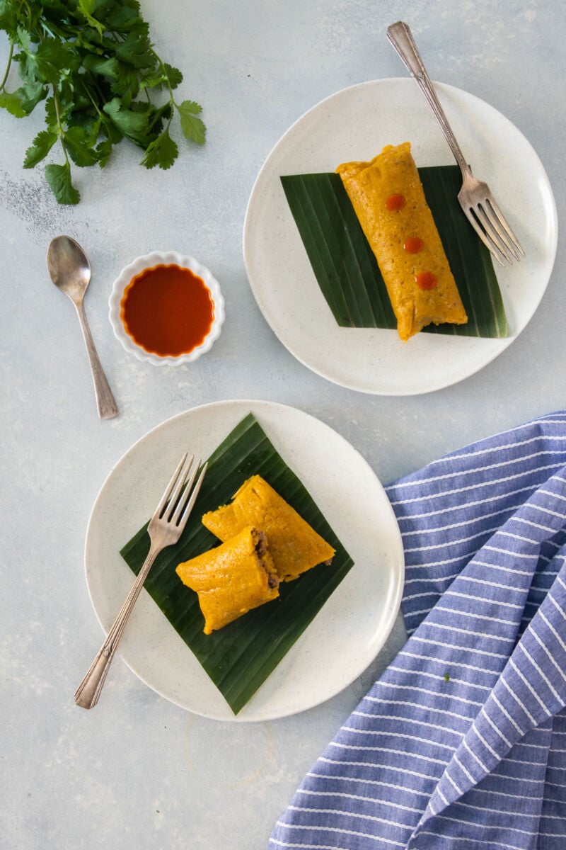 Two pasteles en hojas served on white plates and hot sauce on the side
