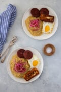 Two plates of mangú with fried eggs, salami, fried cheese, and pickled red onions.