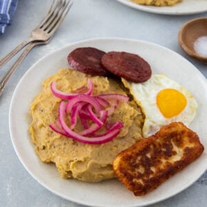 Mangú with fried egg, salami, fried cheese, and pickled red onions on a white plate.