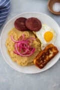 Mangú with fried egg, salami, fried cheese, and pickled red onions on a white plate.