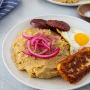 Mangú with fried egg, salami, fried cheese, and pickled red onions on a white plate.
