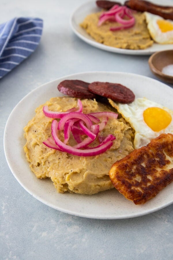 Mangú with fried egg, salami, fried cheese, and pickled red onions on a white plate.