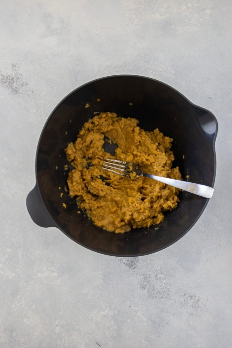 Un bol mezclador negro que contiene plátanos machacados (mangú), con un tenedor en su interior, mostrando una textura suave y machacada.