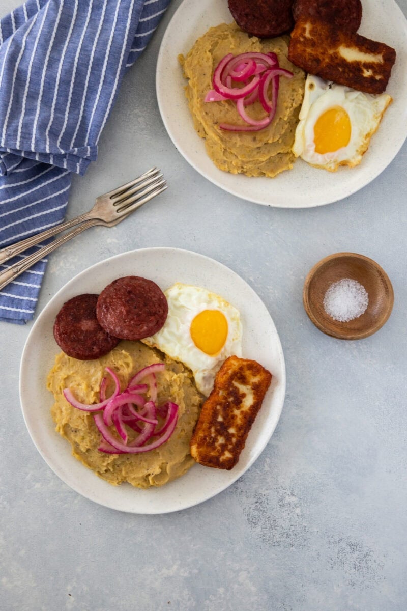 Dos platos de mangú con huevos fritos, salami, queso frito y cebolla roja encurtida.