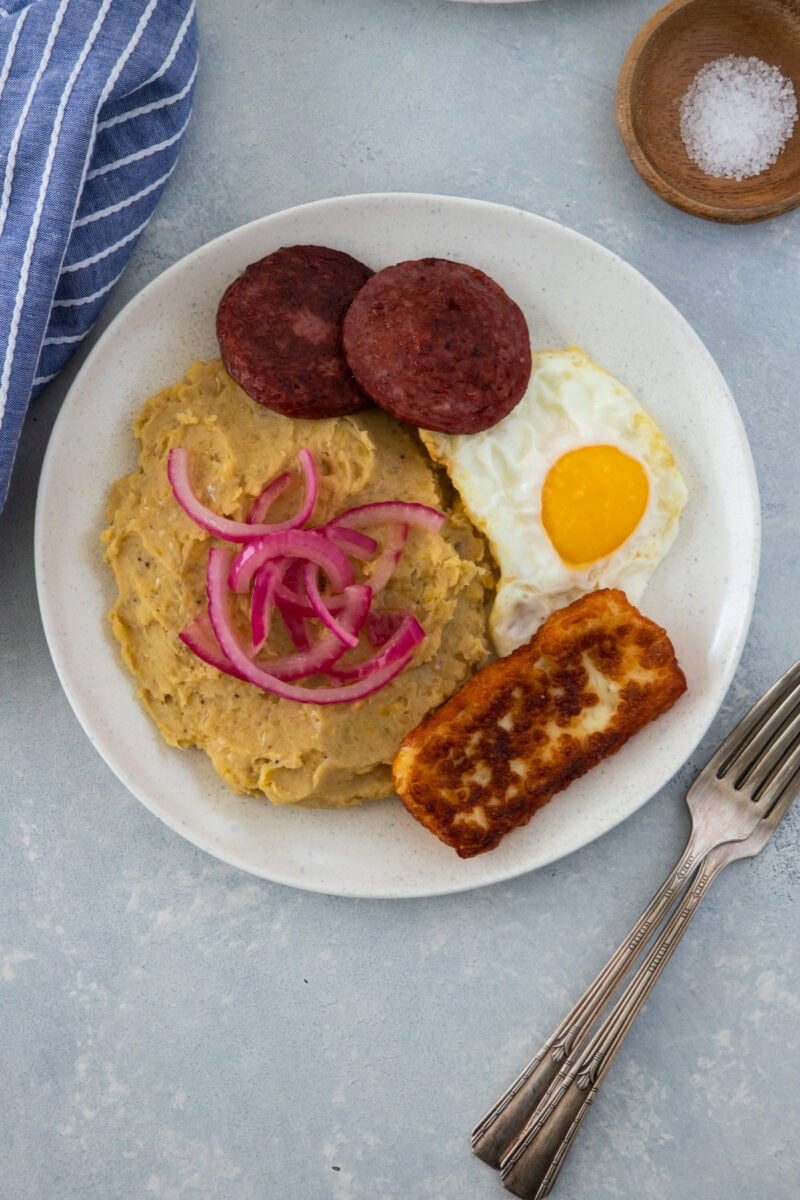 Mangú con huevo frito, salami, queso frito y cebolla roja encurtida en un plato blanco.