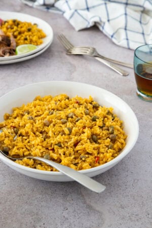 A white deep bowl filled with moro de guandules con coco (Dominican rice with pigeon peas and coconut milk), served with a spoon. In the background, shredded meat, rice, and a lime wedge are visible.