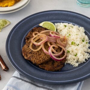 A beef steak topped with onions, served with white rice and a lime wedge, ready to eat.