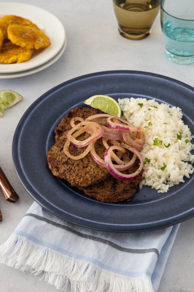 A beef steak topped with onions, served with white rice and a lime wedge, ready to eat.