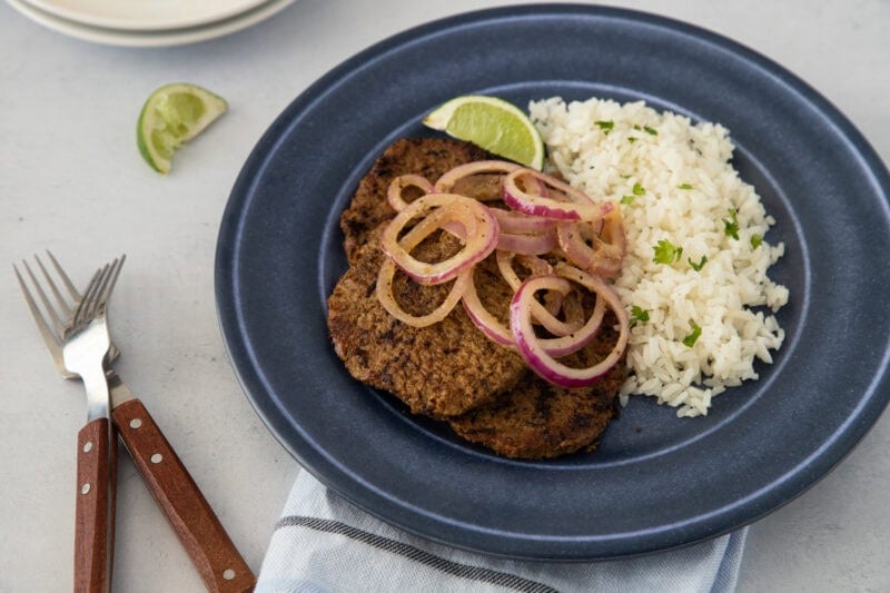 A beef steak topped with onions, served with white rice and a lime wedge, ready to eat.