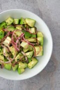 A fresh avocado salad with red onion slices and chopped herbs, served in a white bowl.