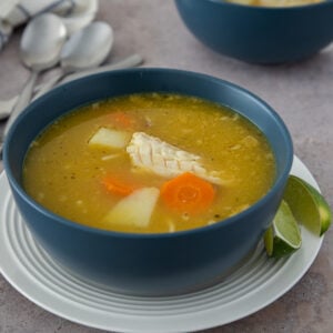 Dominican chicken soup with yuca, carrots, and tender chicken, served in a blue bowl with lime wedges.